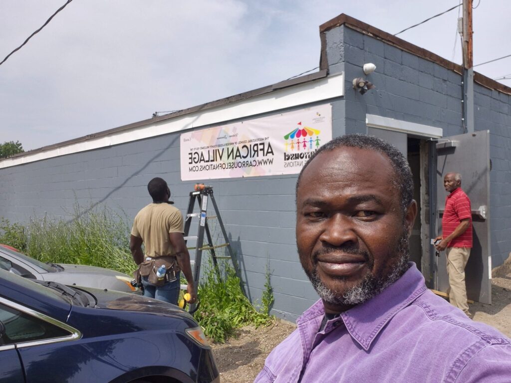 A man standing outside the venue, the African Village banner on the wall behind him.