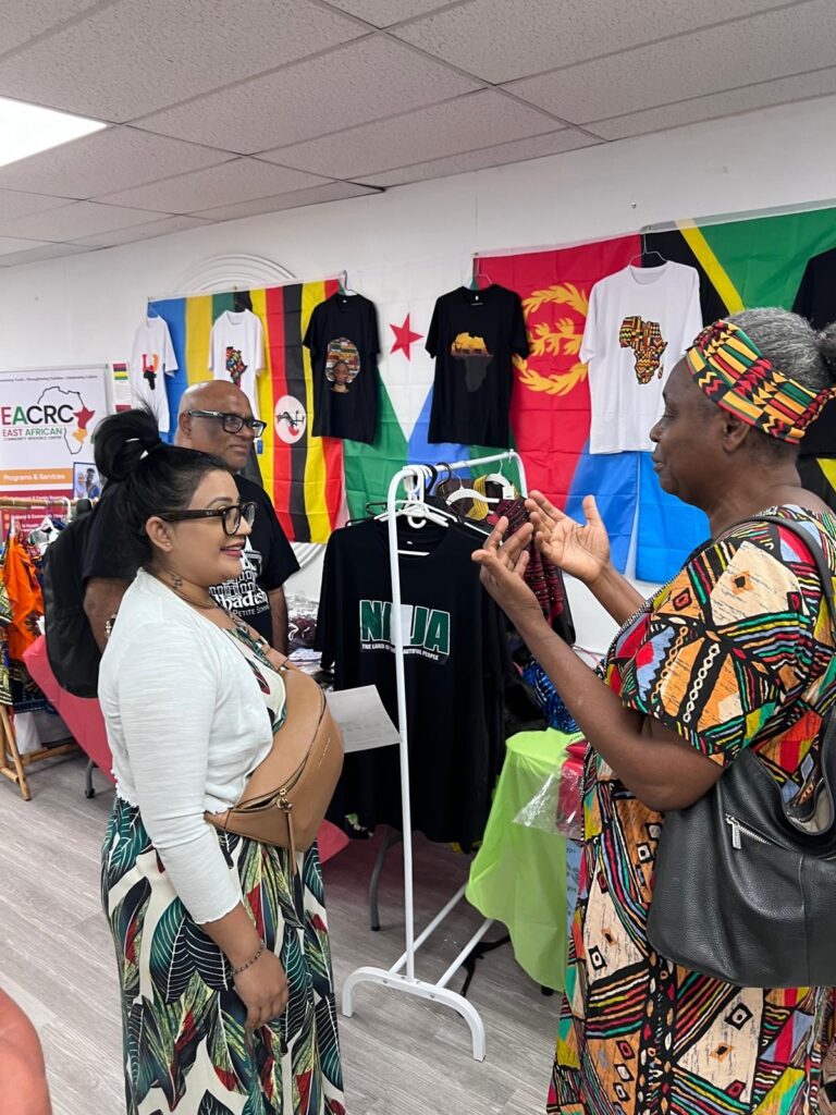 Two visitors talking beside a clothing rail at the BCCEEP Centre, with printed t-shirts hung on the wall above and flags pinned behind them