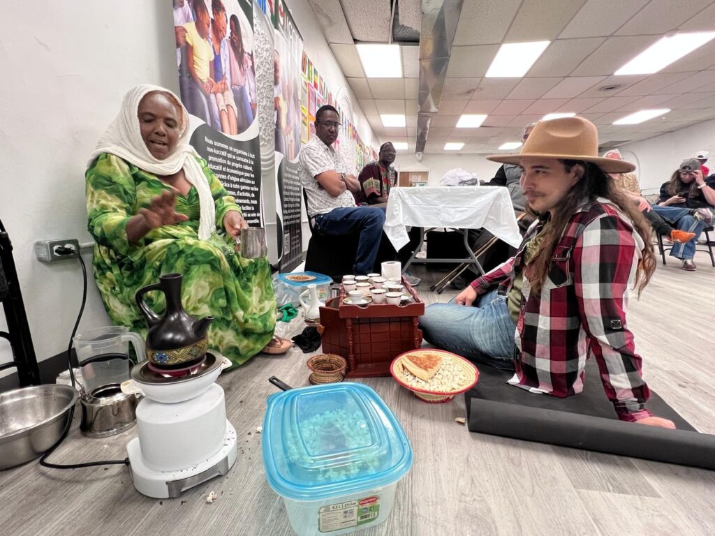 A woman in a green dress and white headscarf pours coffee from a clay pot at the BCCEEP Centre, with a visitor in a wide-brimmed hat seated on the floor beside her and flag bunting along the wall behind