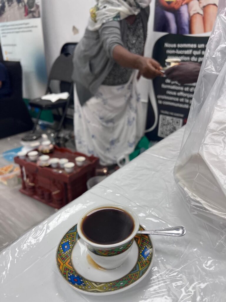 A filled coffee cup on a brightly patterned saucer on the serving table at the BCCEEP Centre, with the server behind it lifting a cup from a wooden stand