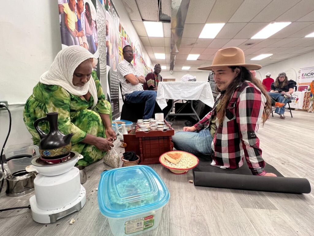 A woman in a green dress tends a clay coffee pot on a small burner on the floor at the BCCEEP Centre, with a tray of cups beside her and guests seated at tables behind