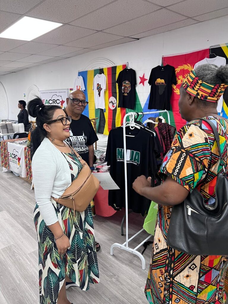 Three visitors in conversation at the BCCEEP Centre in front of a rail of printed shirts, with a display table and a roll-up banner along the far wall