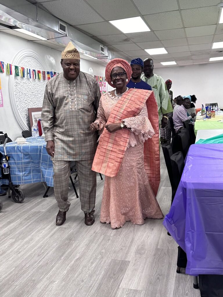 A couple standing together at BCCEEP Hall, one in a gold patterned tunic and cap, the other in a pink lace dress and head tie.