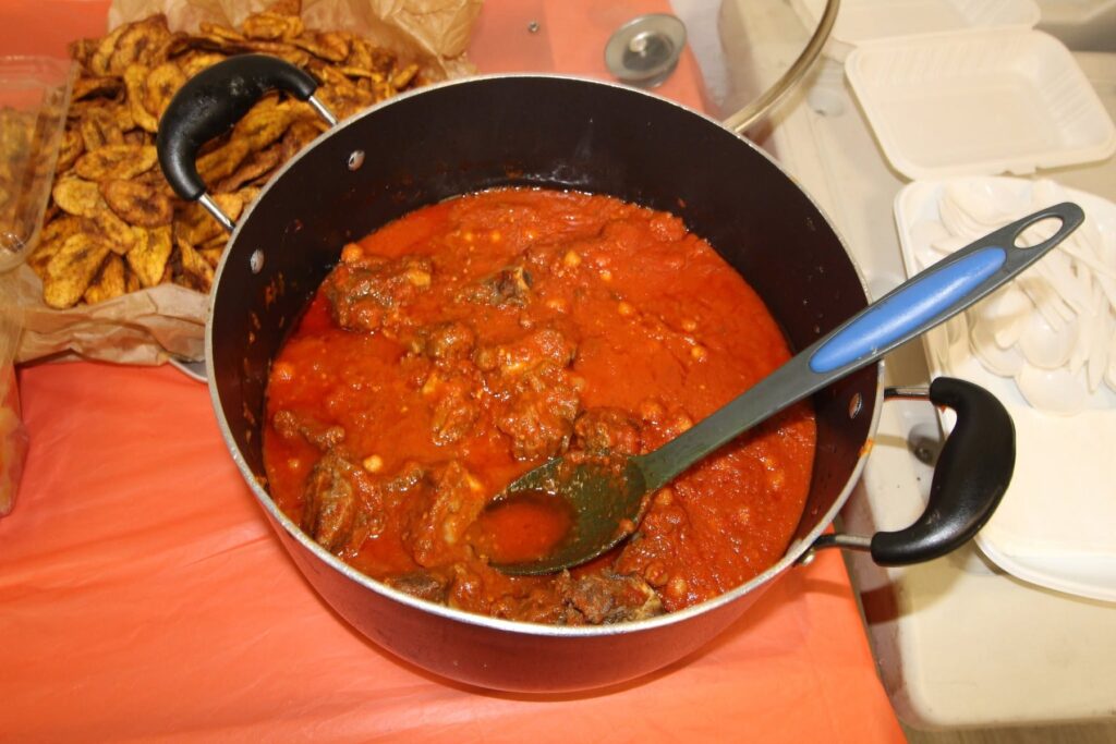 A large pot of red stew with a serving spoon on an orange tablecloth, fried plantain in a dish beside it.