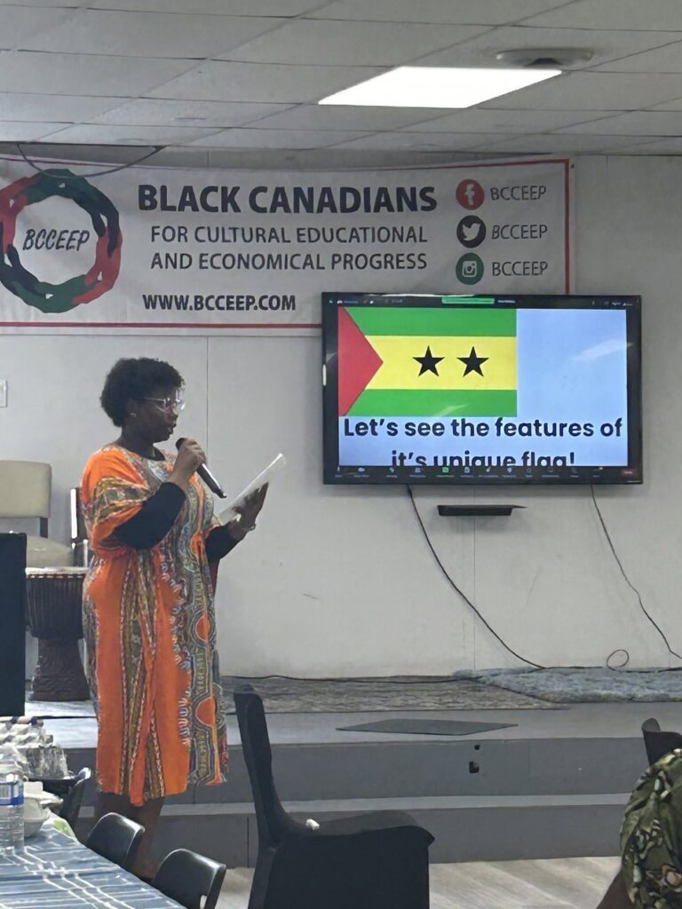 A woman speaking into a microphone beside a wall screen showing a flag, standing in front of the BCCEEP banner.