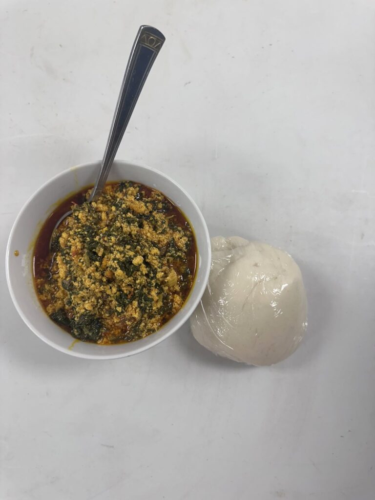 A bowl of green soup with a spoon, served with a smooth white ball of pounded dough alongside.