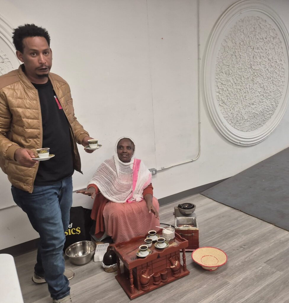 A man standing over a low table set with small cups and a metal bowl at BCCEEP Hall, a seated guest beside him.