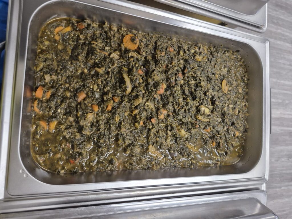 A deep metal tray of cooked greens and vegetables on the serving table at BCCEEP Hall.