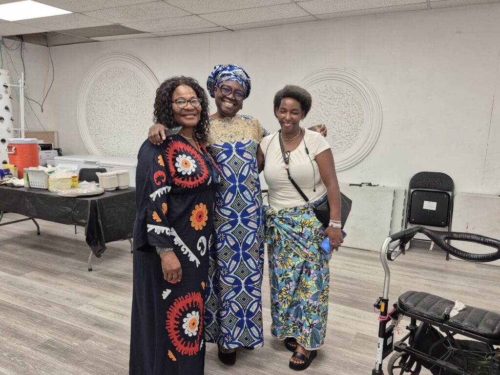 Three guests standing together in bright patterned dress at BCCEEP Hall, a walker and chairs behind them.
