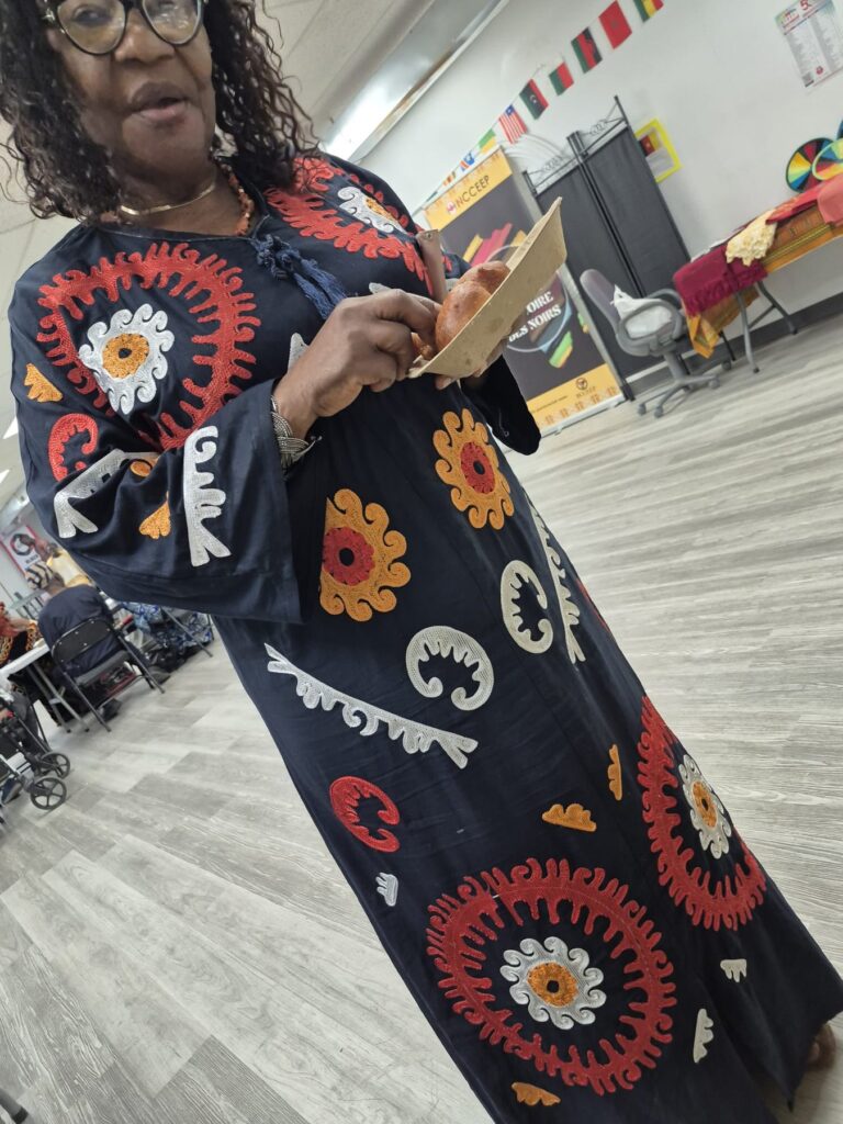 A guest in a long dress printed with large sunflowers standing on the plank floor at BCCEEP Hall, flag bunting on the wall behind.