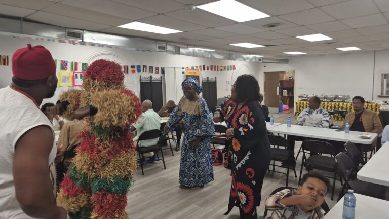 A person in a full raffia costume greeting guests at the BCCEEP Centre, with people seated at tables behind