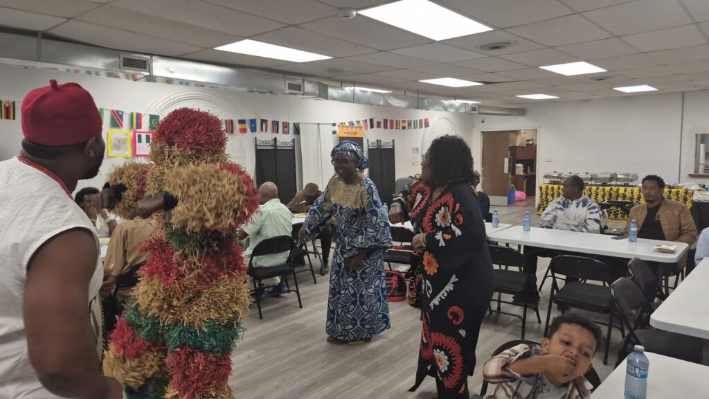 A performer in a layered raffia and fabric costume with a carved mask moving between the tables at BCCEEP Hall.