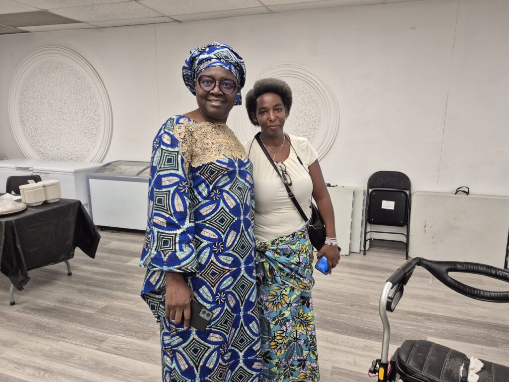 Two guests in blue and green patterned wrappers and head ties standing together in front of the plaster medallions at BCCEEP Hall.