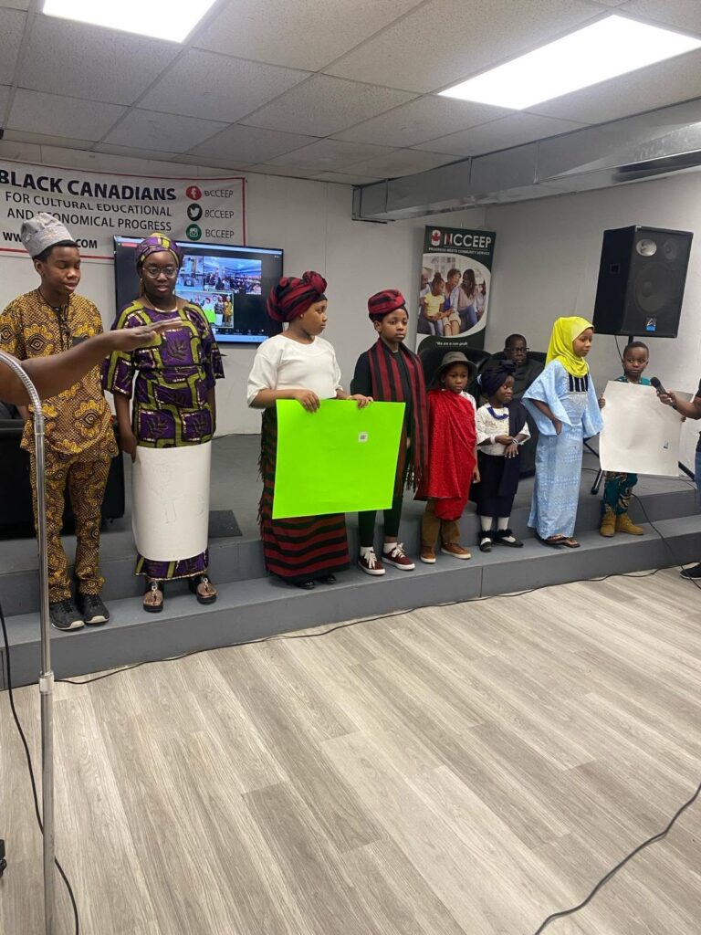 Children standing on the low stage holding up coloured boards while an adult speaks beside them.