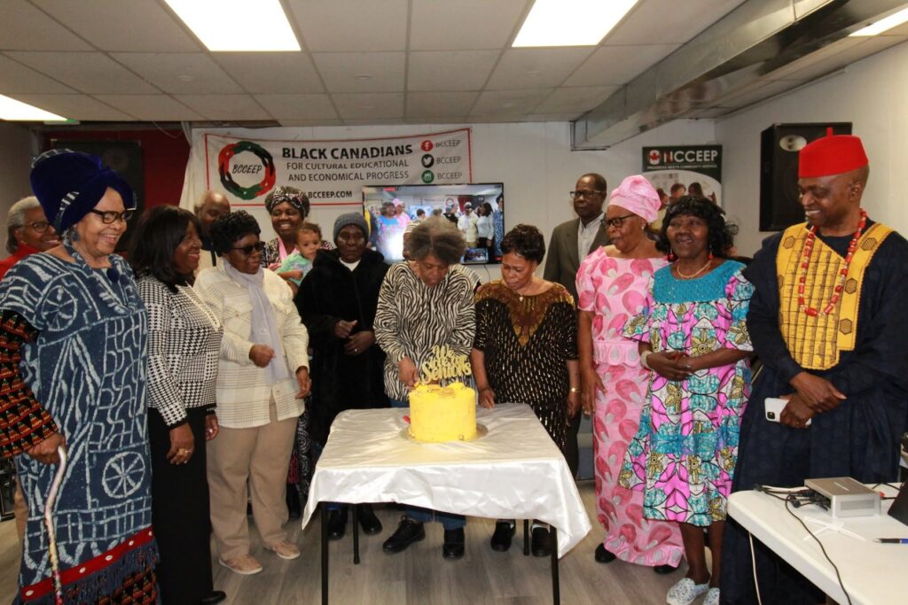 Guests gathered around a yellow cake on a white cloth, one of them cutting the first slice.
