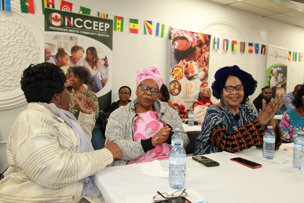 Three guests seated at a table in front of a banner hung with small flags, one raising a hand as she talks.