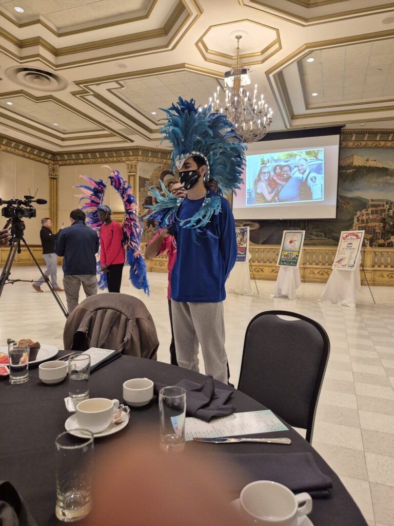 A performer in a tall blue feathered headdress standing beside the presentation screen at the Ciociaro Club.