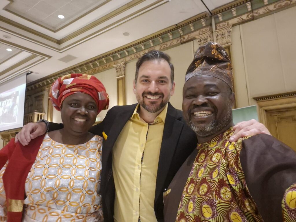 Three guests standing close together at the Ciociaro Club, one in a red head tie and patterned wrapper.