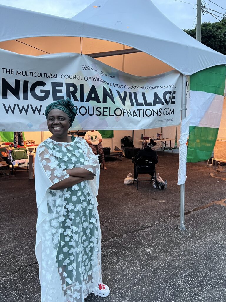 A woman in a pale patterned gown standing and smiling beneath the Nigerian Village banner at the marquee entrance on Walker Road in Windsor