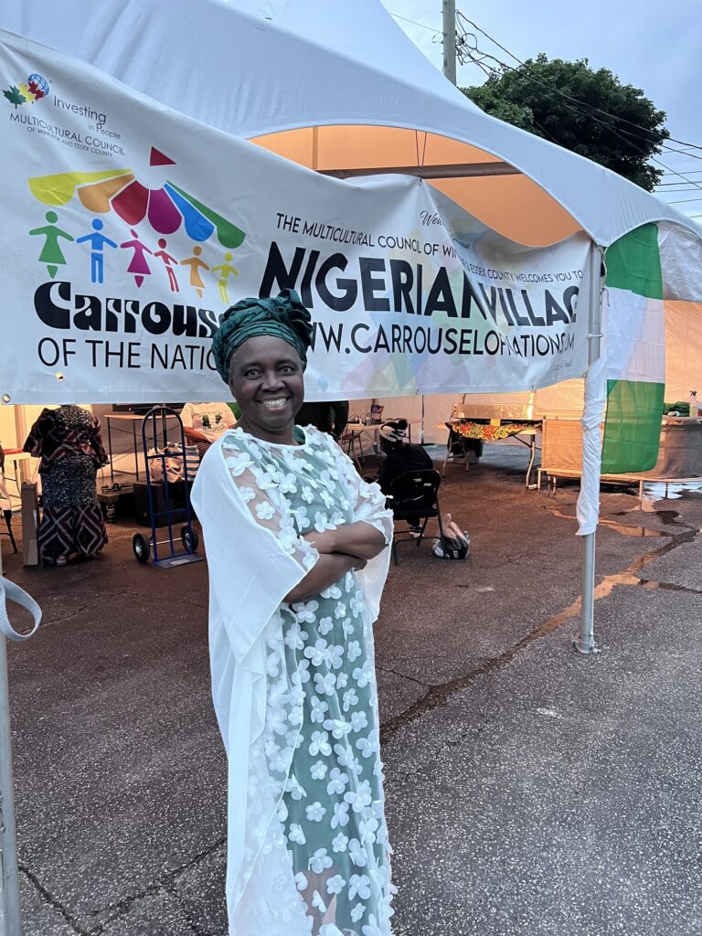 A woman in a pale printed gown smiling beneath the Carrousel of the Nations and Nigerian Village banners at the marquee entrance in Windsor
