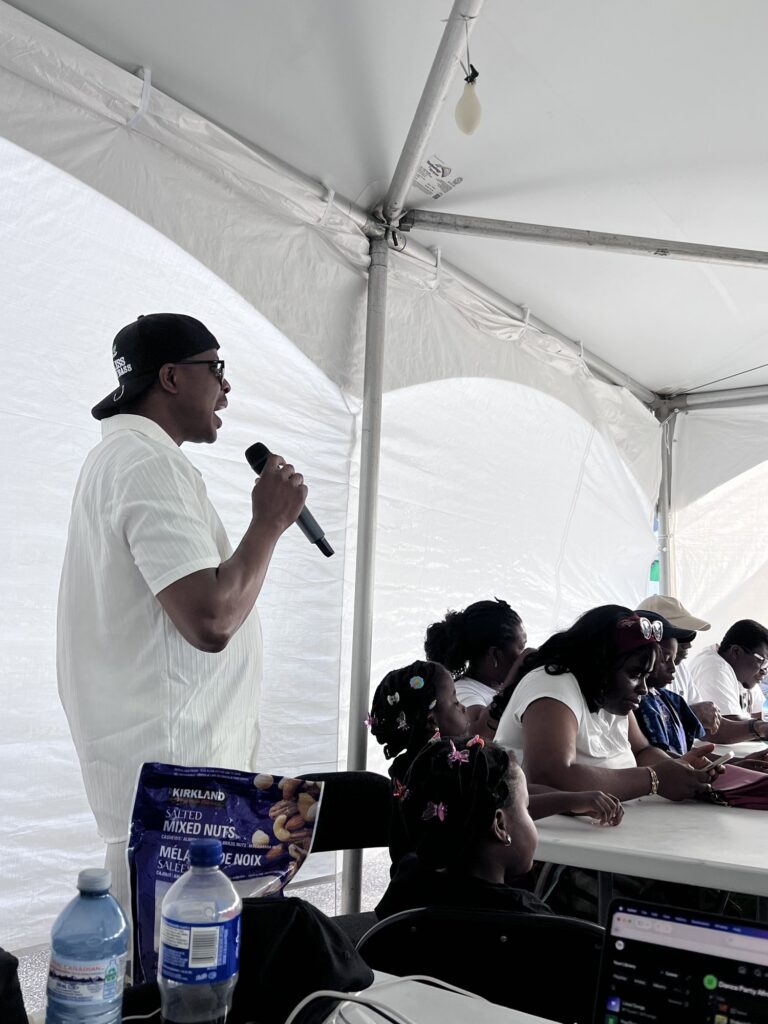 A man in a white shirt and cap holding a microphone while children sit together at the front tables under the marquee at the Nigerian Village