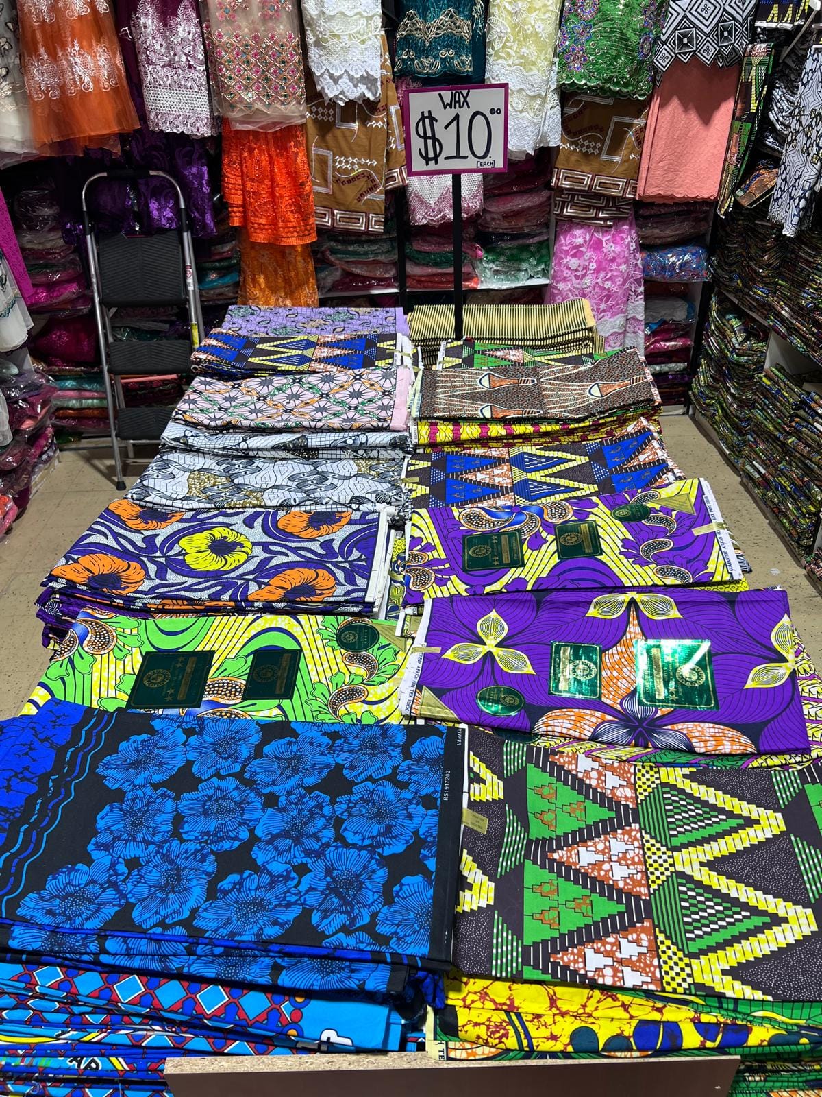 Folded printed fabrics in blue, green and orange laid out across a vendor table at the Nigerian Village, with clothing racks and a ten dollar price sign behind