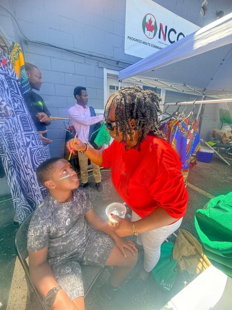 A face painter brushing green and white stripes onto a boy's cheek beside a clothing rack at the Nigerian Village on Walker Road in Windsor