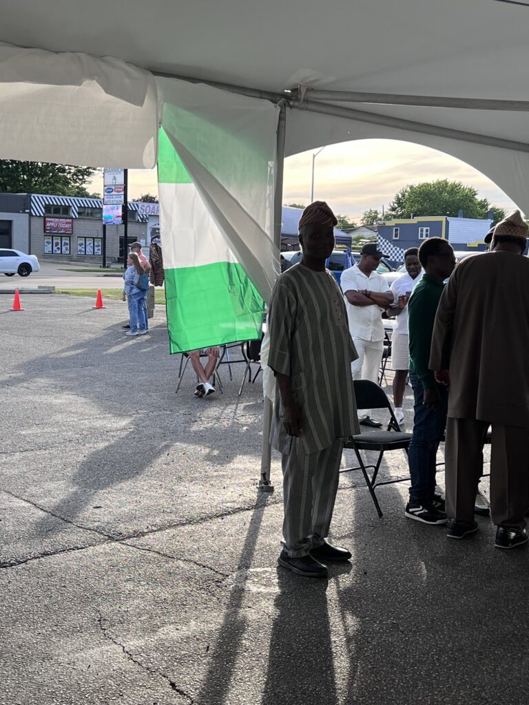 Guests in long pale tunics standing beside a green and white flag hung at the open edge of the marquee at the Nigerian Village