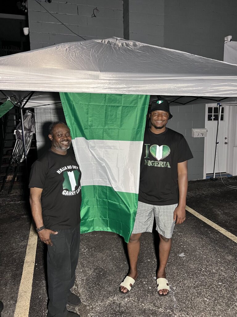 Two men holding a large green and white flag open between them in the parking lot behind the marquee at the Nigerian Village on Walker Road