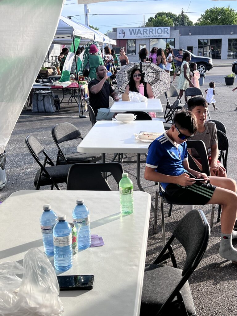 A boy in a blue jersey seated at a table with a plate of food while the market crowd passes behind him at the Nigerian Village