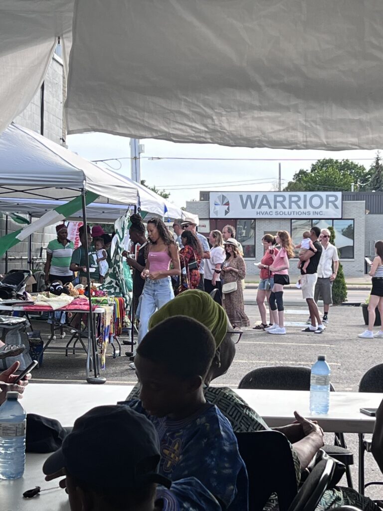 Visitors walking the row of vendor tables at the Nigerian Village on Walker Road, with the Warrior storefront across the street behind them
