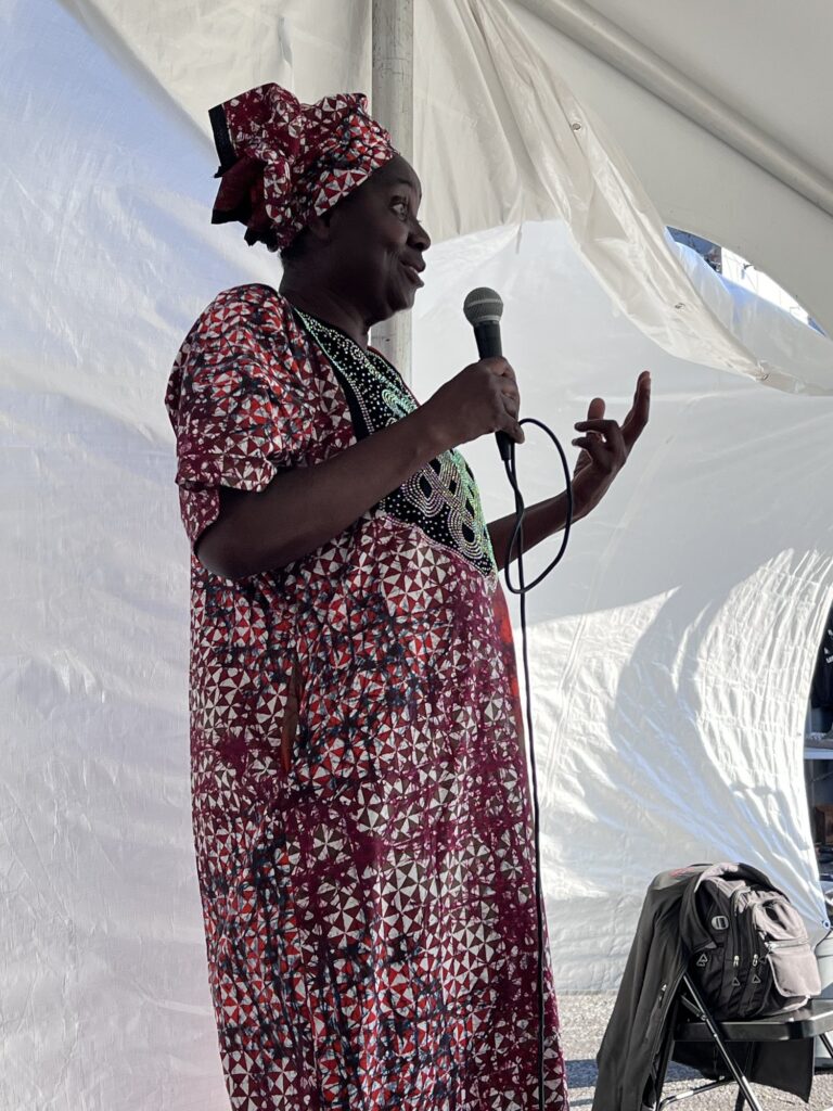 A woman in a red and white printed dress and matching head wrap speaking into a microphone under the marquee at the Nigerian Village