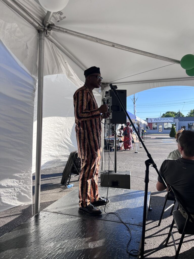 A man in a brown striped tunic and cap addressing the room from the stage end of the marquee at the Nigerian Village on Walker Road