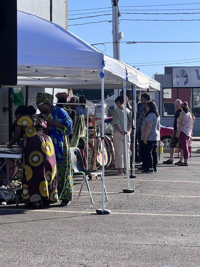 Vendor tables under an open-sided tent at the Nigerian Village, with a guest in a yellow and green patterned wrapper looking over the goods on display