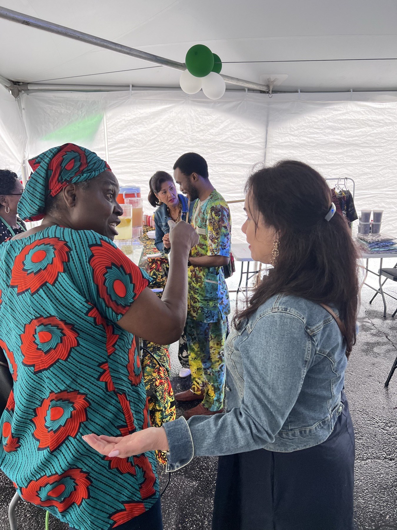 Two guests in conversation under the marquee at the Nigerian Village, one wearing a red and teal patterned dress with a matching head wrap