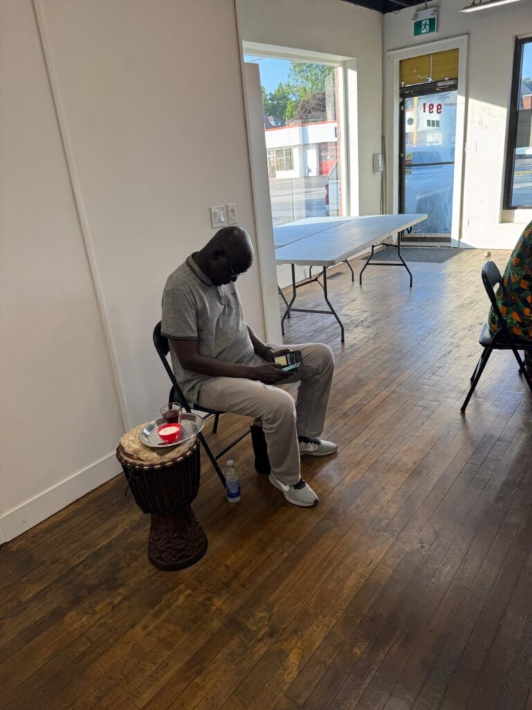 A man seated on a folding chair with a drum standing on the floor in front of him.